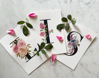 May include: Three watercolor paintings of floral letters. The letters are L, I, and S. Each letter is surrounded by pink, white, and purple flowers.