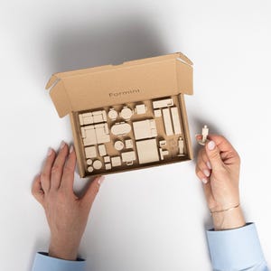 Może przedstawiać: Miniaturowy zestaw drewnianych mebli w kartonowym pudełku. Pudełko jest otwarte, ukazując różne meble w małej skali. Osoba trzyma małą drewnianą figurkę. Pudełko jest oznaczone "Formini".