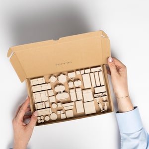 Op de afbeelding: Een kartonnen doos met het label "Formini" bevat een verzameling miniatuur houten meubels. De set bevat banken, tafels, bedden en andere meubels, allemaal in een natuurlijke houten afwerking. De doos wordt door twee handen vastgehouden.