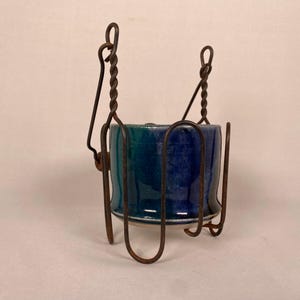 May include: A blue and green ceramic planter with a white rim sits in a rusty metal hanging planter. The planter has a twisted wire design.