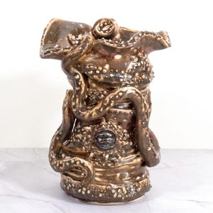 May include: A ceramic vase shaped like an octopus with brown and white glaze. The vase has a textured surface and a wide opening at the top.