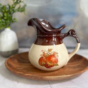 May include: A brown and cream ceramic pitcher with a fruit design. The pitcher has a wide mouth and a handle. It is sitting on a wooden tray.