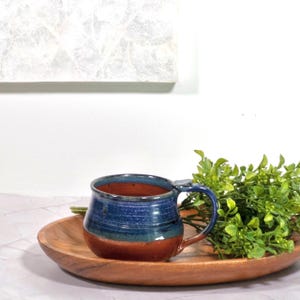 May include: A handmade ceramic mug with a blue and brown glaze. The mug has a rounded shape with a handle and sits on a wooden tray. The mug is approximately 4 inches tall and 3 inches in diameter.