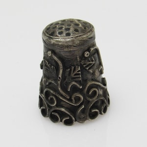 May include: A silver thimble featuring an intricate design of swirls and leaves. The top of the thimble has a small hole for threading.