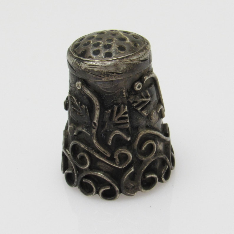 May include: A silver thimble featuring an intricate design of swirls and leaves. The top of the thimble has a small hole for threading.