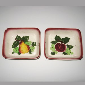 May include: Two square ceramic dishes with red trim. One dish features a painted pear with green leaves and grapes. The other dish features a painted apple with green leaves and grapes.