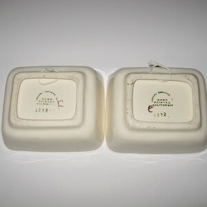 May include: Two white ceramic square dishes with rounded edges. The bottom of each dish has a green and gold sticker that reads "Jacona Ceramics, Hand Painted, California, 1232."