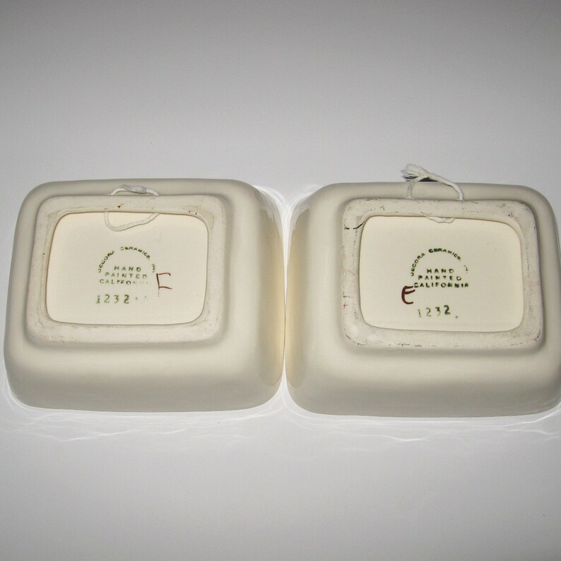 May include: Two white ceramic square dishes with rounded edges. The bottom of each dish has a green and gold sticker that reads "Jacona Ceramics, Hand Painted, California, 1232."