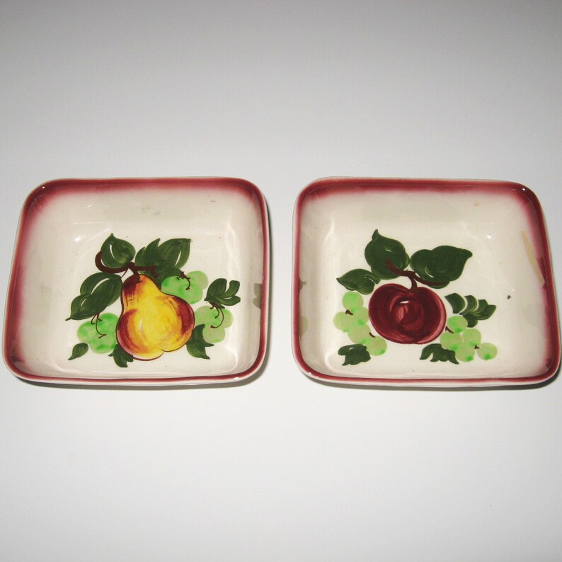 May include: Two small, square ceramic dishes with a red rim. One dish features a painted pear with green leaves and grapes. The other dish features a painted apple with green leaves and grapes.
