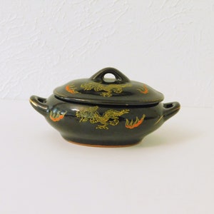 May include: A small black ceramic serving dish with a lid. The dish is decorated with gold and red dragons and flames.