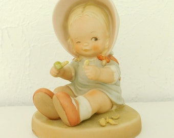 May include: A porcelain figurine of a little girl wearing a pink hat and a blue dress. She is sitting on a base and holding a small green object in her hand. She is winking and smiling.