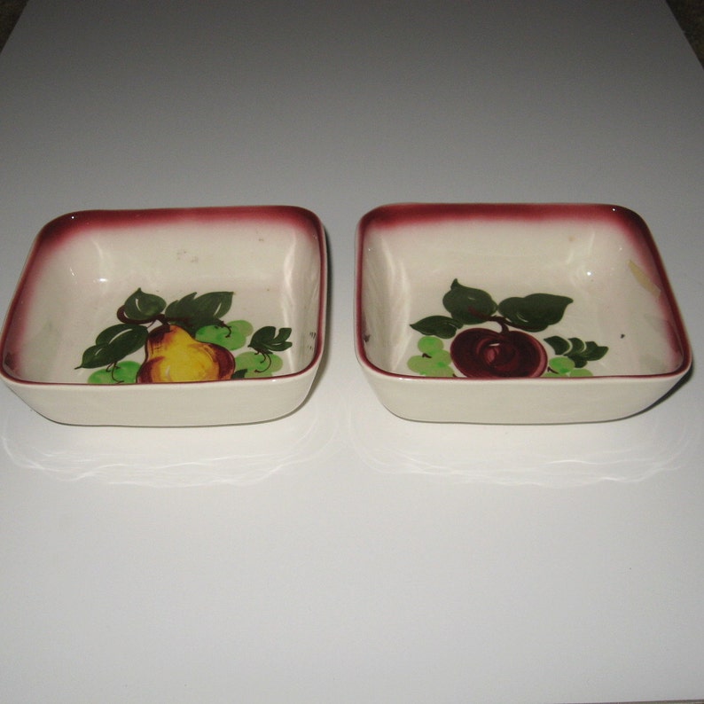 May include: Two square ceramic bowls with a white background and a burgundy rim. One bowl features a painted pear with green leaves and grapes, while the other bowl features a painted apple with green leaves and grapes.