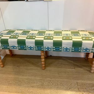 May include: A rectangular bench with a green and white checkered pattern seat. The bench has turned wooden legs. The seat is upholstered with a woven fabric. The legs are a light brown color.
