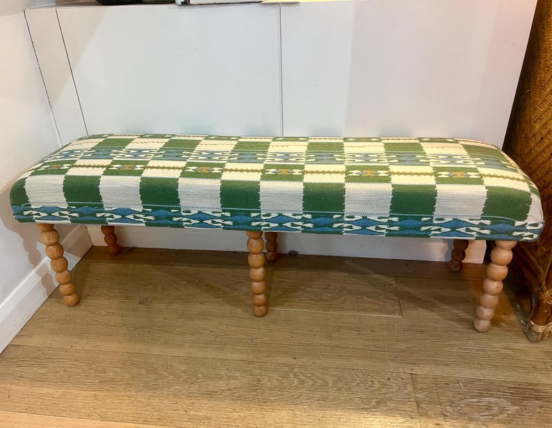 May include: A rectangular bench with a green and white checkered pattern seat. The bench has turned wooden legs. The seat is upholstered with a woven fabric. The legs are a light brown color.