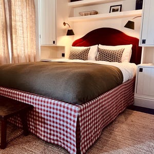 May include: A bedroom with a red velvet headboard, white pillows, and a bed skirt with a red and white checkered pattern. The bed is covered with a brown blanket. The room has white walls, shelves with books, and sconce lighting.
