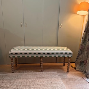 Puede incluir: Banco rectangular con asiento a cuadros verdes y blancos. El banco tiene patas de madera torneadas. El fondo presenta un armario verde claro y una lámpara de pie con una pantalla naranja. Una alfombra está en primer plano.