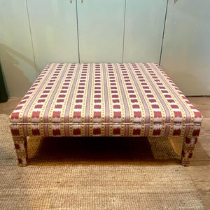 Pode incluir: Um pufe quadrado com um tampo de tecido estampado. O tecido apresenta listras verticais em vermelho, creme e dourado. O pufe tem um perfil baixo e é suportado por quatro pernas curtas e quadradas.