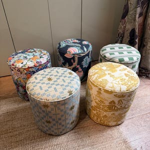 Puede incluir: Cinco otomanas cilíndricas con diversos estampados de tela. Una tiene un estampado floral colorido, otra un diseño floral oscuro, otra rayas verdes y blancas, otra un patrón geométrico de estrellas azul claro y otra un estampado de paisaje toile amarillo.
