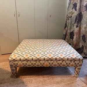Op de afbeelding: Een vierkante poef met een crèmekleurige stof met een herhalend patroon van oranje sterren en bruine geometrische vormen. De poef heeft korte, vierkante poten en staat op een geweven vloerkleed. Afmetingen: ongeveer 76 cm x 76 cm.