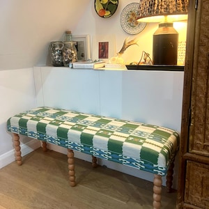 May include: A rectangular bench with a green, white, and blue patterned cushion. The bench has four wooden, turned legs. The bench is in a room with a wooden floor and white walls. Decorative plates hang on the wall.