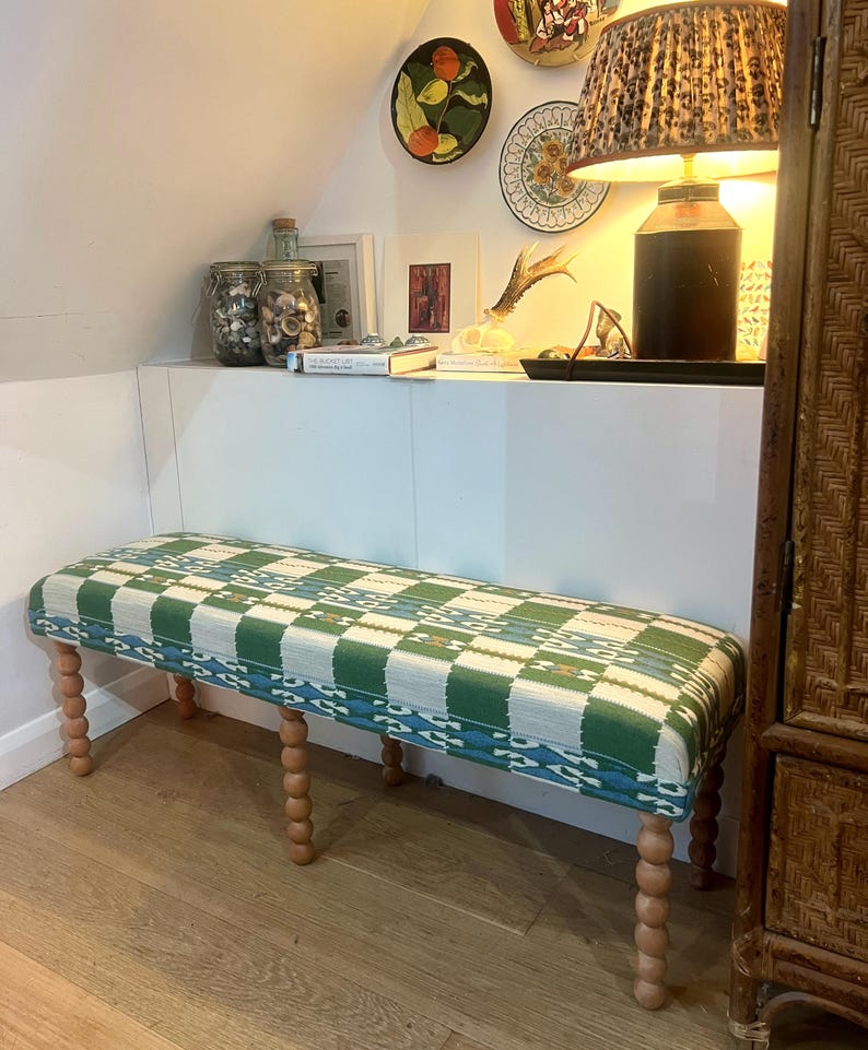 May include: A rectangular bench with a green, white, and blue patterned cushion. The bench has four wooden, turned legs. The bench is in a room with a wooden floor and white walls. Decorative plates hang on the wall.