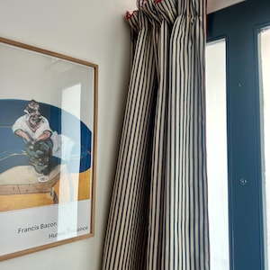 May include: A pair of striped curtains with a red trim hanging in front of a window. The curtains are made of a cream and navy blue striped fabric. The window is black and has a glass pane.