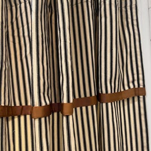 May include: Cream-colored curtains with vertical black stripes. The curtains are adorned with a brown ribbon detail near the bottom. Gold-colored curtain rings are visible at the top. The curtains are made of a textured fabric.