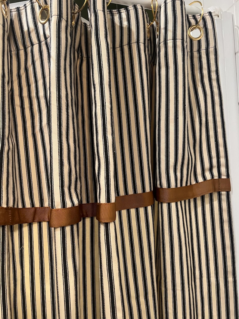May include: Cream-colored curtains with vertical black stripes. The curtains are adorned with a brown ribbon detail near the bottom. Gold-colored curtain rings are visible at the top. The curtains are made of a textured fabric.
