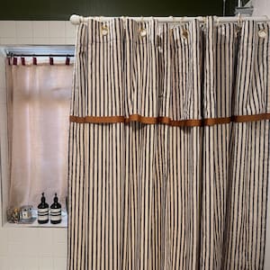 May include: A striped shower curtain with vertical black and off-white stripes. The curtain has a brown band across the top and is hung with gold-colored rings. A window with a striped curtain is visible to the left.