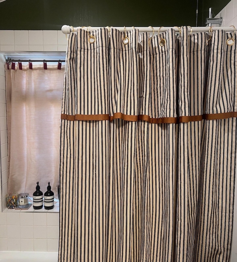 May include: A striped shower curtain with vertical black and off-white stripes. The curtain has a brown band across the top and is hung with gold-colored rings. A window with a striped curtain is visible to the left.