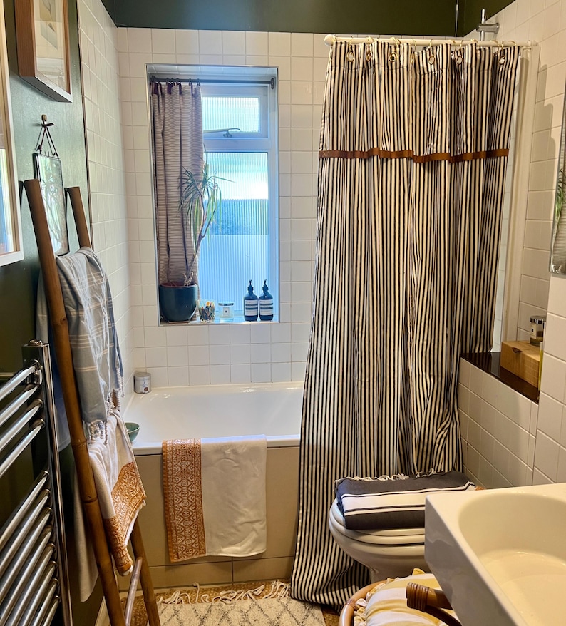May include: A bathroom interior featuring a white bathtub, a striped shower curtain, and a bamboo towel rack. The shower curtain has vertical black and white stripes with brown accents. A window with a sheer curtain is visible.