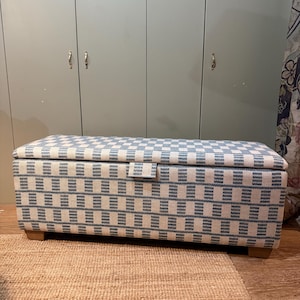 Puede incluir: Un otomán de almacenamiento rectangular con un patrón de cuadros azules y blancos. El otomán tiene una tapa con bisagras y se apoya en cuatro pequeños pies de madera. El fondo presenta un armario verde claro y una alfombra beige.