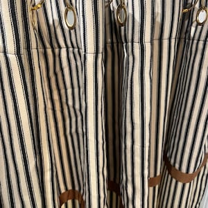 May include: Cream-colored and black striped shower curtains with gold-colored shower curtain rings. The curtains have a brown ribbon trim along the bottom edge. The vertical stripes create a classic, timeless look for a bathroom.
