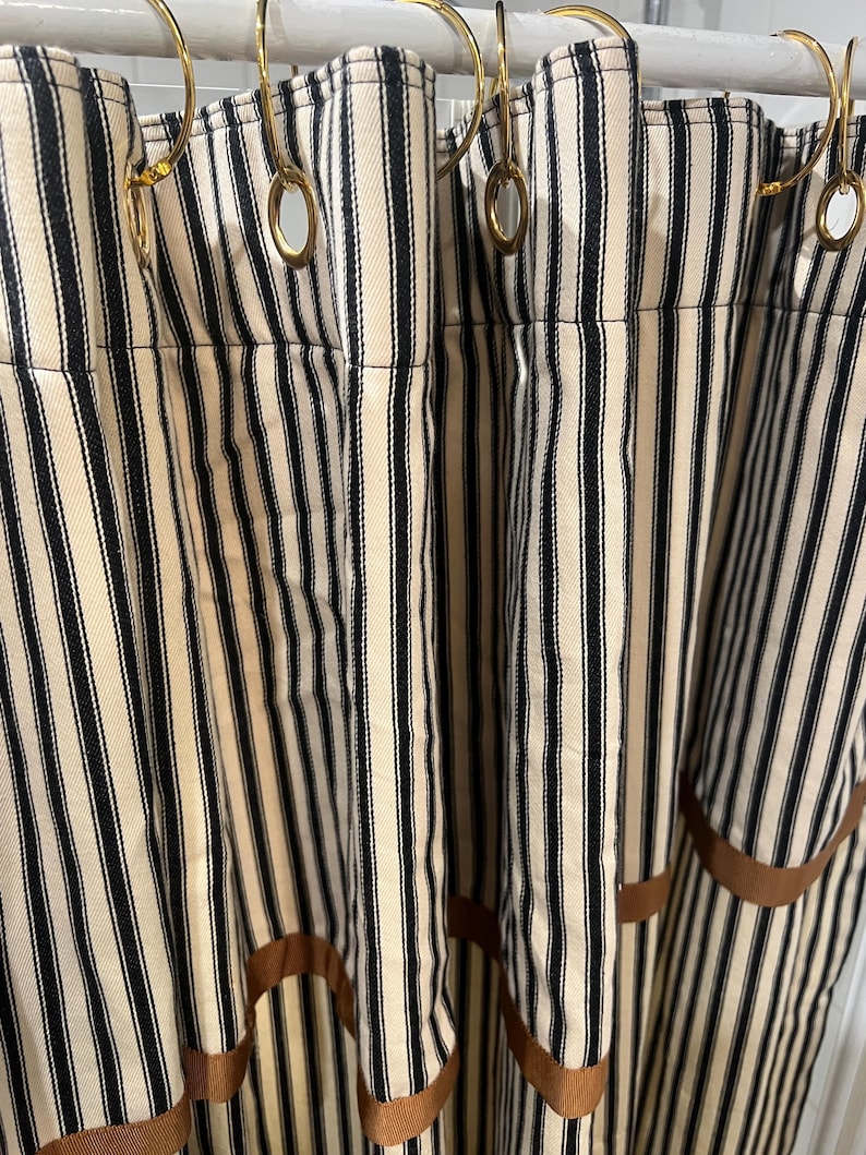 May include: Cream-colored and black striped shower curtains with gold-colored shower curtain rings. The curtains have a brown ribbon trim along the bottom edge. The vertical stripes create a classic, timeless look for a bathroom.