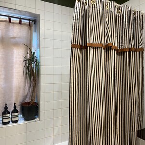 May include: A bathroom scene featuring a striped shower curtain with brown accents. The curtain has vertical black and white stripes. A window with a sheer curtain and a potted plant is visible. Two dark bottles and a candle sit on the window sill.