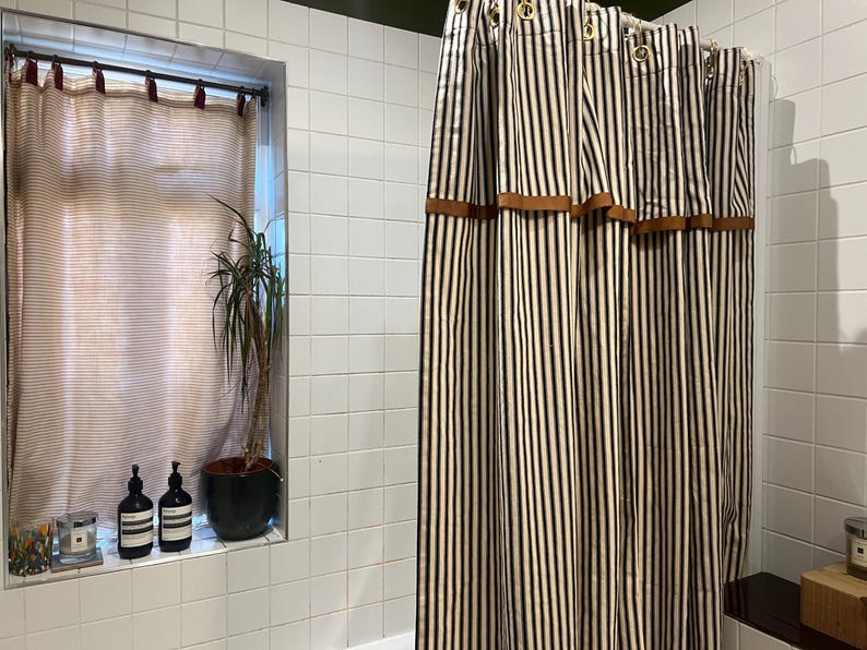 May include: A bathroom scene featuring a striped shower curtain with brown accents. The curtain has vertical black and white stripes. A window with a sheer curtain and a potted plant is visible. Two dark bottles and a candle sit on the window sill.