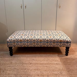 Puf otomano Ashby da Bespoke Studio, com acabamento em debrum e opção de pés torneados.