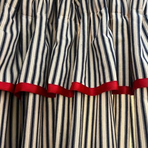 May include: Close-up of striped curtains. The curtains feature vertical navy blue and white stripes with a red ribbon trim along the bottom edge. The fabric is gathered at the top, creating soft folds.