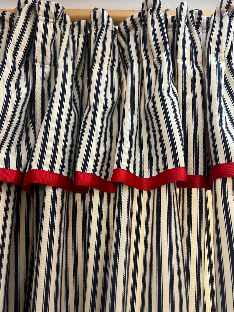 May include: Close-up of striped curtains. The curtains feature vertical navy blue and white stripes with a red ribbon trim along the bottom edge. The fabric is gathered at the top, creating soft folds.