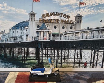 May include: A blue and white vintage scooter model with a black seat and chrome accents. The scooter is in front of a backdrop of a pier with the words "Brighton Palace Pier" on the building.