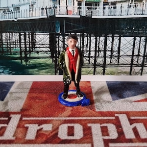 Könnte beinhalten: Eine Miniaturfigur einer Person, die eine grüne Jacke, eine rote Weste und eine Krawatte trägt. Die Figur steht auf einem blauen und roten Sockel mit einem weißen Kreis in der Mitte. Die Figur steht vor einem Foto eines Piers.