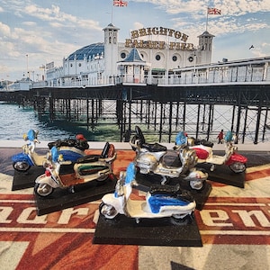 Könnte beinhalten: Vier Miniatur-Modelle von Rollern, die in verschiedenen Farben lackiert sind, darunter Weiß, Blau, Rot und Grün. Die Roller werden auf schwarzen Sockeln vor einem Hintergrund aus einer rot-weiß-blauen gemusterten Oberfläche präsentiert. Der Hintergrund zeigt einen Pier mit dem sichtbaren Text "Brighton Palace Pier".