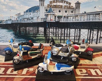 May include: A collection of five miniature metal scooter figurines, each mounted on a black rectangular base. The scooters feature various color schemes including blue, white, red, and silver, with detailed handlebars, seats, and wheels, displayed against a backdrop of Brighton Palace Pier.