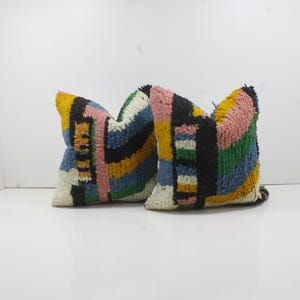 以下が含まれることがあります： カラフルな幾何学模様の正方形の装飾用枕2つ。枕は、黄色、ピンク、青、黒、緑、白などの色の組み合わせが特徴です。枕は、質感のある素材で作られています。
