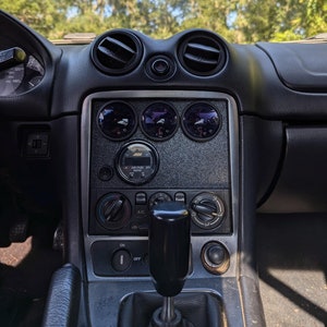 May include: Interior view of a car dashboard with black trim and air vents. The center console features gauges and controls, including a gear shift and climate control knobs. The background shows trees and a blue sky.