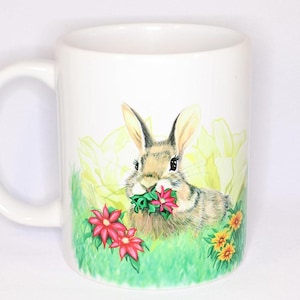 Peut inclure: Mug en céramique blanche avec une illustration à l'aquarelle d'un lapin entouré de fleurs et d'herbe. Le lapin tient des fleurs dans sa bouche. La tasse a une anse incurvée et une finition lisse et brillante.