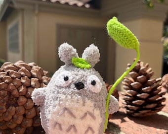 May include: A handmade crochet Totoro figure in shades of gray, with a white belly and black eyes. It holds a green leaf on a stem. The figure is displayed on a wooden base, with pine cones in the background.