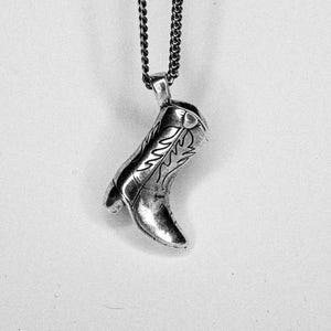 Puede incluir: Un collar con colgante plateado que presenta un dije detallado de bota de vaquero. El dije de la bota tiene patrones decorativos grabados y está suspendido de una cadena oscura. Un accesorio de temática occidental.