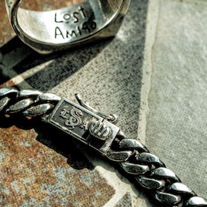 May include: A silver chain bracelet with a clasp that has the words "The Saga" engraved on it. The bracelet is lying on a textured surface.