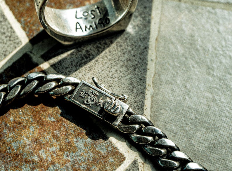 May include: A silver chain bracelet with a clasp that has the words "The Saga" engraved on it. The bracelet is lying on a textured surface.